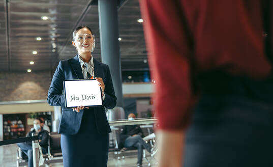 Frau mit Gesichtsschutz, die am Flughafenterminal steht und ein Namensschild hält, um einen Reisenden zu empfangen. - JLPSF12624