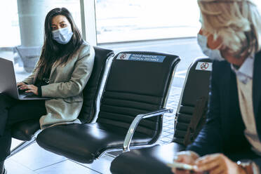 Businesswoman using laptop and talking with a male passenger while waiting at airport terminal. Business travelers wearing protective face masks waiting for their flights. - JLPSF12629