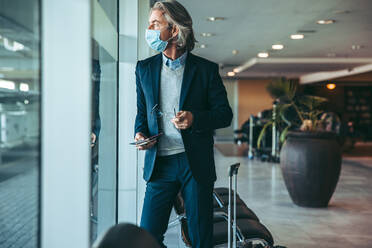 Businessman standing at airport lounge with suitcase and looking outside the window. Man with face mask waiting for his flight in airport terminal. - JLPSF12630