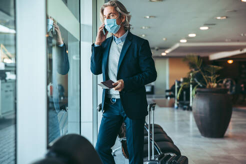 Geschäftsmann mit Gesichtsmaske, der in der Wartelounge eines Flughafens steht und ein Mobiltelefon benutzt. Männlicher Reisender, der im Flughafenterminal auf seinen Flug wartet. - JLPSF12631
