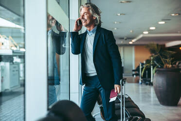 Geschäftsmann steht in der Flughafenlounge und spricht mit seinem Handy. Männlicher Geschäftsreisender am Flughafen wartet auf den Flug. - JLPSF12632