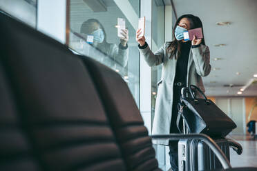 Woman wearing face mask video calling with her cell phone and showing her boarding pass. Female traveler having a video call at airport waiting lounge during pandemic. - JLPSF12638