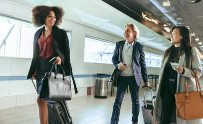 Lächelnde Geschäftsleute, die auf Geschäftsreise sind und Koffer tragen, während sie durch den Flughafengang gehen. - JLPSF12644