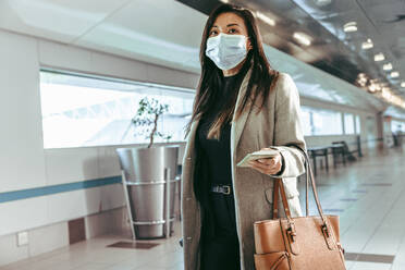 Masked woman travelling through a plane. Female flight passenger walking towards boarding gate with luggage. - JLPSF12656