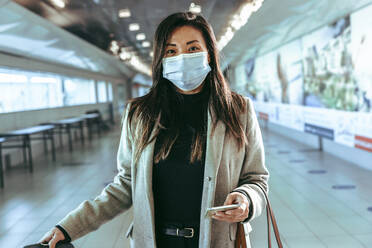 Portrait of a woman traveler wearing protective face mask standing at airport terminal hallway. Female passenger with mask looking at camera. - JLPSF12658