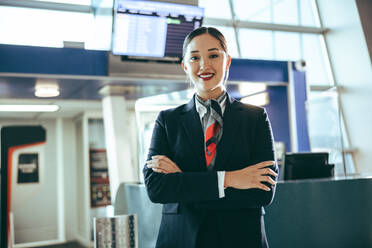 Porträt einer glücklichen jungen Frau, die auf einem Flughafen arbeitet. Flugbegleiterin, die mit verschränkten Armen auf einem internationalen Flughafen am Flugsteig steht. - JLPSF12675