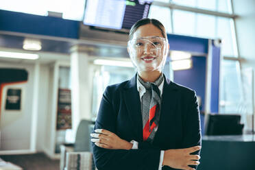 Porträt einer weiblichen Flugbegleiterin bei der Arbeit während einer Pandemie: Frau mit Gesichtsschutz, die mit verschränkten Armen im Flughafenterminal steht. - JLPSF12676