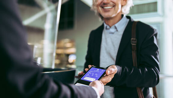 Cropped shot of a male passenger check in at airport terminal using digital flight ticket. Businessman showing plane boarding pass to staff on mobile phone. - JLPSF12682