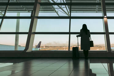 Rückansicht einer weiblichen Reisenden, die durch das Fenster schaut, während sie auf ihren Flug am Flughafen wartet. Frau wartet am Flughafen-Terminal. - JLPSF12689