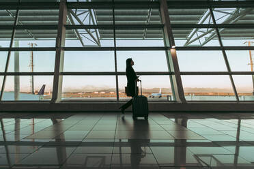 Silhouette einer weiblichen Flugpassagierin mit Gepäck, die im Flughafenterminal spazieren geht. Eine Reisende geht vor einem großen Fenster im Flughafenterminal. - JLPSF12691