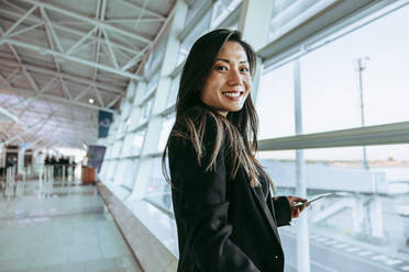 Porträt einer glücklichen Frau, die im Flughafenterminal steht und auf den Flug wartet. Weibliche Reisende in der Flughafenlounge, die am Fenster steht. - JLPSF12693