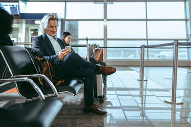 Älterer Mann, der in der Flughafenlounge auf seinen Flug wartet. Geschäftsmann, der auf einer Bank sitzt und ein Mobiltelefon im Wartebereich des Flughafens benutzt. - JLPSF12698