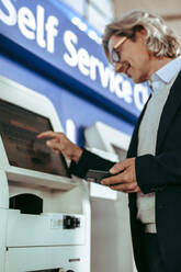 Niedriger Blickwinkel eines Mannes, der den Selbstbedienungs-Check-in-Automaten am Flughafen benutzt. Älterer Geschäftsmann beim Einchecken am Flughafen. - JLPSF12718