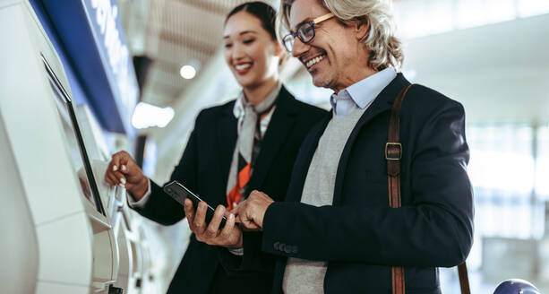 Glücklicher Geschäftsmann beim Self-Check-in am Automaten mit Flugbegleiterin im Flughafenterminal. Passagier am Self-Service-Check-in-Automaten mit weiblichem Flughafenpersonal. - JLPSF12721