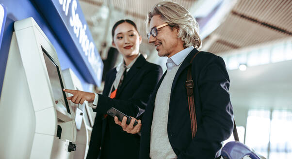 Eine weibliche Flugbegleiterin hilft einem männlichen Reisenden mit dem Self-Check-in-Automaten im Flughafenterminal während einer Pandemie. Selbstbedienungs-Check-in-Schalter auf einem internationalen Flughafen. - JLPSF12722