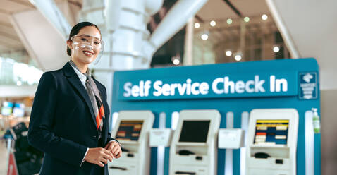 Glückliche junge Frau, die während einer Pandemie als Bodenpersonal am Flughafen arbeitet. Weibliche Flughafenmitarbeiterin, die vor einem Selbstbedienungs-Check-in-Automaten am Flughafen steht und in die Kamera schaut und lächelt. - JLPSF12729