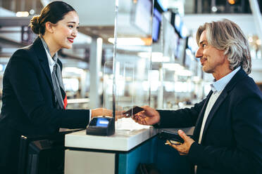 Reifer Geschäftsmann beim Einchecken am Check-in-Schalter einer Fluggesellschaft auf einem internationalen Flughafen. Geschäftsreisender, der dem Flughafenpersonal seinen Pass übergibt, wobei eine Glaswand zwischen ihnen steht. - JLPSF12730