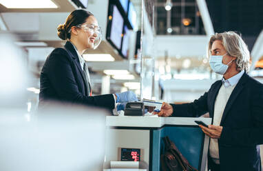 Flugbegleiter mit Gesichtsschutz, der einem Passagier am Check-in-Schalter hilft. Eine Frau, die am Flughafen arbeitet und einen Reisenden am Flugsteig betreut. - JLPSF12735