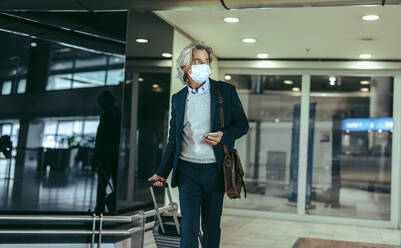 Businessman wearing face mask pulling suitcase out of airport terminal. Business traveler arriving at from flight during pandemic. - JLPSF12741