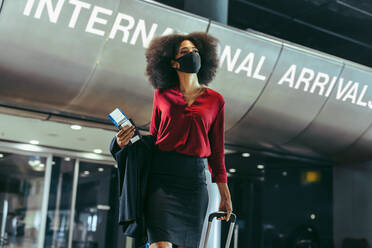 Female business traveler wearing face mask with her luggage walking in front of international arrival gate in airport. Businesswoman walking through airport terminal. - JLPSF12743