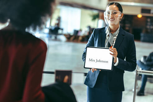 Weiblicher Fahrer mit Gesichtsschutz, der am Flughafenterminal steht und ein Namensschild hält, um einen Reisenden zu empfangen. Chauffeur mit einem Schild am Ankunftsterminal. - JLPSF12747