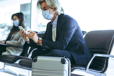 Businessman sitting on chair at airport and using mobile phone. Business traveler waiting for this flight at airport during pandemic. - JLPSF12752