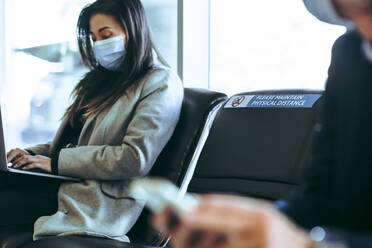 Businesswoman sitting at airport terminal working on laptop. International business traveler wearing face mask waiting for her flight during pandemic. - JLPSF12753