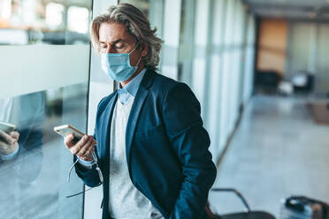 Geschäftsmann mit Gesichtsmaske, der im Wartebereich eines Flughafens steht und sein Mobiltelefon benutzt. Männlicher Reisender, der im Abflugbereich eines Flughafens auf seinen Flug wartet. - JLPSF12754