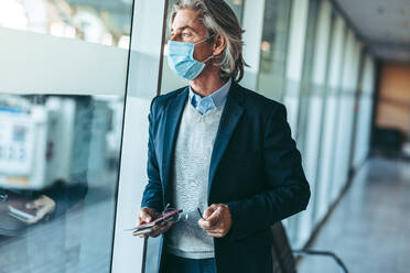 Business traveler wearing face mask standing at airport waiting lounge and looking outside the window. Businessman waiting for his flight during coronavirus outbreak. - JLPSF12755