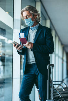 Mann mit Gesichtsmaske im Wartebereich eines Flughafens, der sein Mobiltelefon benutzt. Reisende, die im Abflugbereich eines Flughafens auf ihren Flug warten. - JLPSF12756