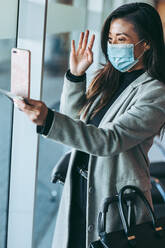 Woman wearing face mask video calling with her cell phone. Female traveler having a video call at airport waiting lounge. - JLPSF12766