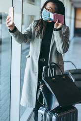 Woman wearing face mask showing her boarding pass on video call while waiting at airport terminal. Female traveler video calling from airport waiting lounge. - JLPSF12767