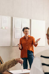 Cheerful businesswoman pitching her idea to her team during a meeting. Happy team leader having a discussion with her colleagues. Group of creative businesspeople working together in an office. - JLPSF12782