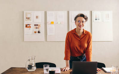 Mature interior designer smiling at the camera while standing behind her desk in her office. Happy businesswoman with eyeglasses working with a laptop in a creative office. - JLPSF12814