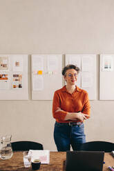 Happy female designer smiling at the camera while standing behind her desk with her arms crossed. Mature businesswoman with eyeglasses working in a creative office. - JLPSF12818