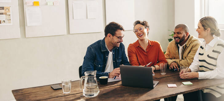 Lächelnde Geschäftsleute, die an einem Laptop in einem kreativen Büro diskutieren. Eine Gruppe glücklicher Geschäftsleute tauscht Ideen aus, während sie gemeinsam an einem neuen Innenarchitekturprojekt arbeiten. - JLPSF12840