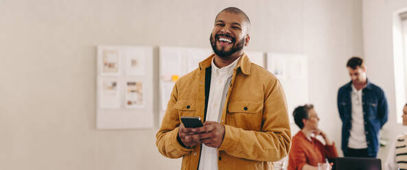 Cheerful businessman with a septum ring smiling at the camera while holding a smartphone. Happy young businessman working in a creative and modern workplace with his colleagues. - JLPSF12844