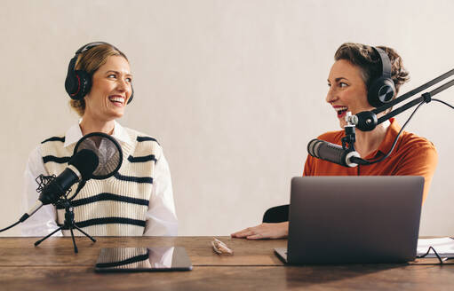 Fröhliche Podcasterinnen unterhalten sich bei einer Audioübertragung in einem Heimstudio. Zwei fröhliche Frauen nehmen einen Internet-Podcast für ihren Social-Media-Kanal auf. - JLPSF12861