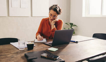 Mature businesswoman writing in her diary while sitting in front of a laptop in her office. Female designer writing down her to-do list while working in a creative office. - JLPSF12869