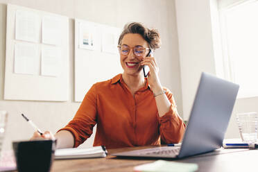 Happy businesswoman speaking on the phone while sitting at her desk with a notebook and a laptop. Smiling interior designer making plans with a client in a creative office. - JLPSF12873