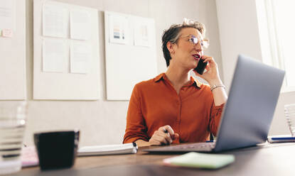 Eine reife Geschäftsfrau telefoniert, während sie mit Notebook und Laptop an ihrem Schreibtisch sitzt. Eine Innenarchitektin schmiedet Pläne mit einem Kunden in einem kreativen Büro. - JLPSF12874