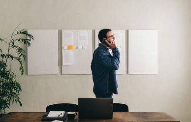 Young businessman talking on the phone while standing behind his desk in a creative office. Businessman communicating with his clients while working remotely. - JLPSF12886