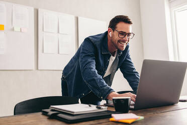 Happy businessman smiling cheerfully while connecting with his team online. Young businessman joining a virtual meeting on a laptop. Businessman with eyeglasses working remotely in a creative office. - JLPSF12890