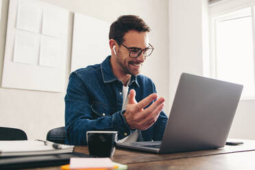 Glücklicher Geschäftsmann, der lächelt, während er mit seinem Team über einen Videoanruf spricht. Fröhlicher Geschäftsmann, der ein Online-Meeting an einem Laptop abhält. Geschäftsmann mit Brille, der aus der Ferne in einem kreativen Büro arbeitet. - JLPSF12892