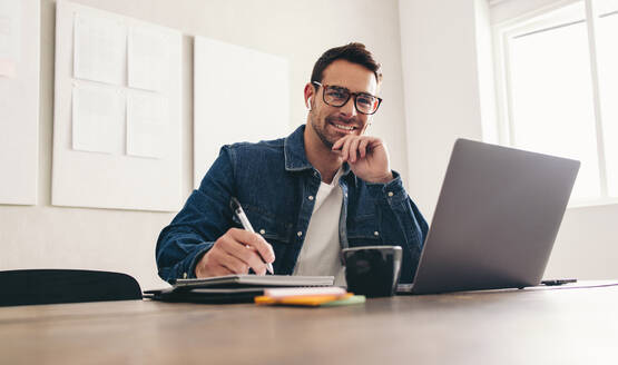 Making notes during a video conference. Happy young businessman smiling at the camera while sitting in an online meeting with a notebook. Cheerful businessman working remotely in a creative office. - JLPSF12893