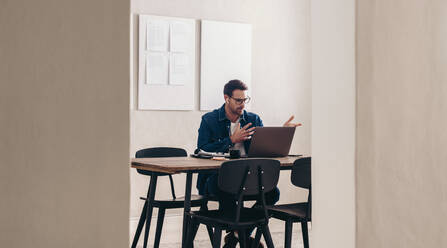 Junger Geschäftsmann, der während einer Videokonferenz mit seinem Team spricht. Geschäftsmann, der ein Online-Meeting auf einem Laptop abhält. Junger Geschäftsmann mit Brille, der aus der Ferne in einem kreativen Büro arbeitet. - JLPSF12894