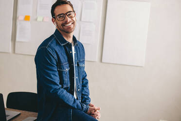 Happy young businessman having a conference call using wireless earbuds. Cheerful businessman smiling while communicating with his team. Businessman with glasses working remotely in a creative office. - JLPSF12896