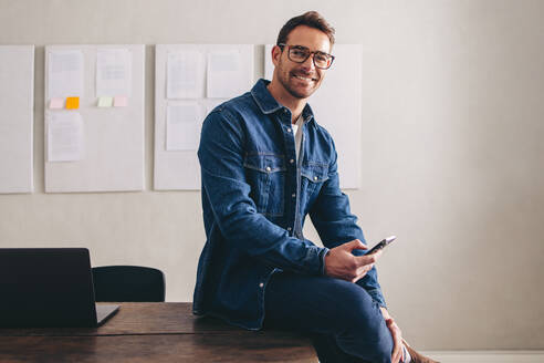 Glücklicher junger Geschäftsmann, der in die Kamera lächelt, während er mit einem Smartphone auf dem Schreibtisch sitzt. Fröhlicher Geschäftsmann mit Brille, der aus der Ferne in einem kreativen Büro arbeitet. - JLPSF12904