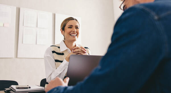 Fröhliche Geschäftsfrau lächelt während eines Treffens mit einem Geschäftspartner in ihrem Büro. Glückliche junge Geschäftsfrau, die eine Zusammenarbeit mit ihrem Geschäftspartner plant. - JLPSF12908