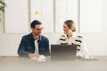 Lächelnde Geschäftsleute, die sich mit einem Laptop auf ein Online-Meeting vorbereiten. Zwei glückliche Geschäftsleute, die sich in einem kreativen Büro mit ihren globalen Kunden verbinden. - JLPSF12915